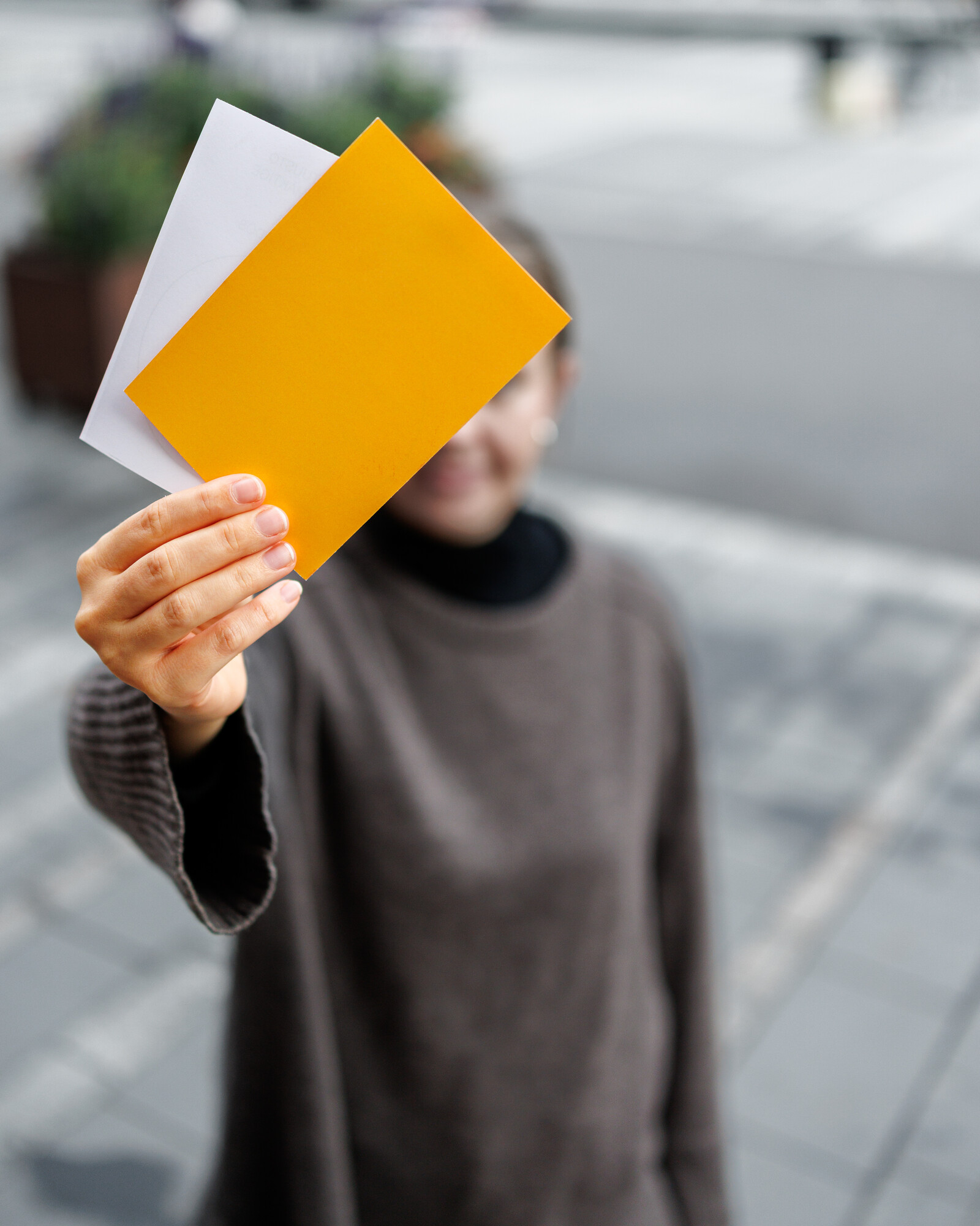 Harmaapaitainen henkilö pitää kädessään kahta paperia, yksi keltainen ja yksi valkoinen, taustalla epätarkka kaupunkimaisema. Paperit peittävät suurimman osan henkilön kasvoista.