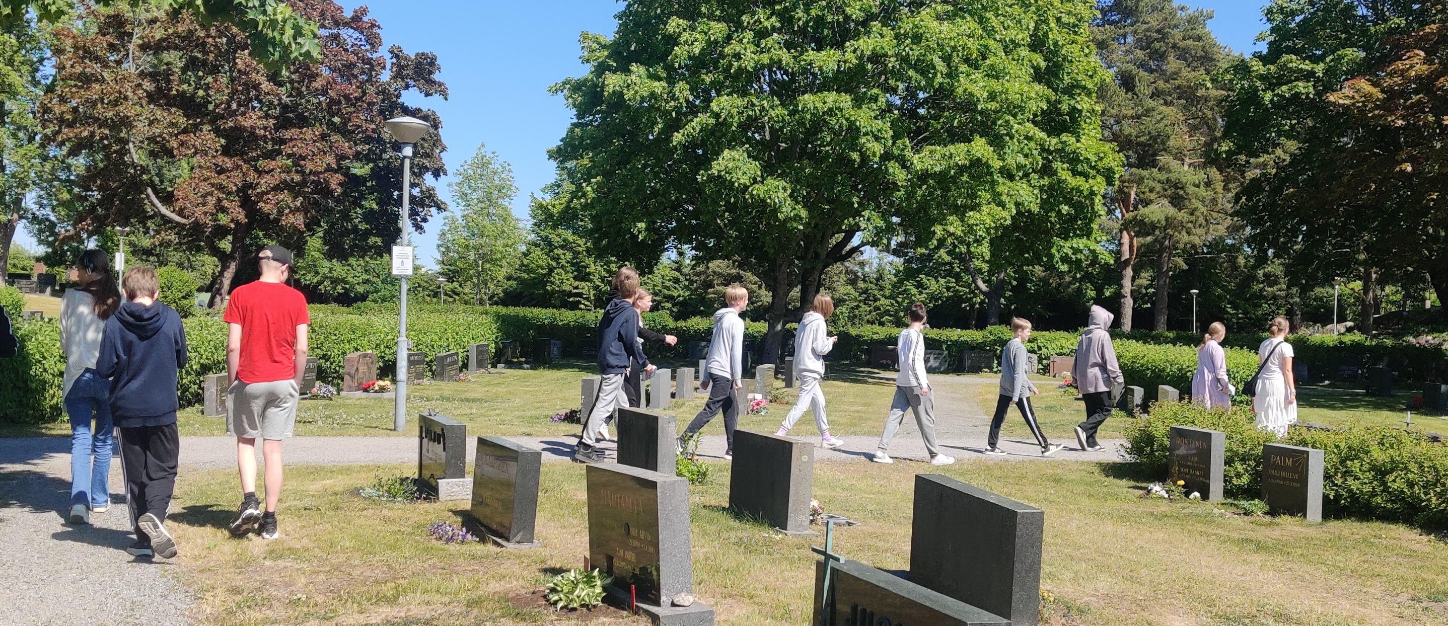 Kymmenkunta nuorta ja kaksi aikuista kävelemässä hautausmaan poikki kesällä.